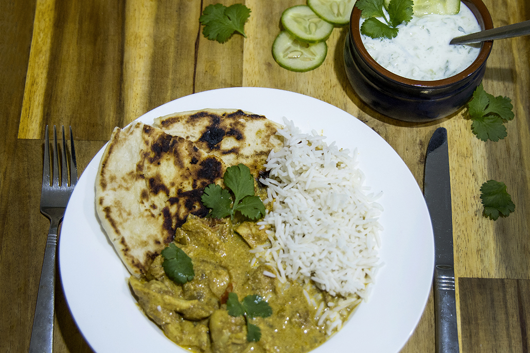 Butter chicken with rice, naan bread and cucumber raita served on a plate.